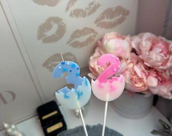 Puede incluir: Dos velas de cumpleaños sobre palitos blancos, una con forma de número tres en azul claro con puntos y grietas azul oscuro, y otra con forma de número dos en rosa con puntos y grietas rosa oscuro, ambas emergiendo de una cáscara de huevo.