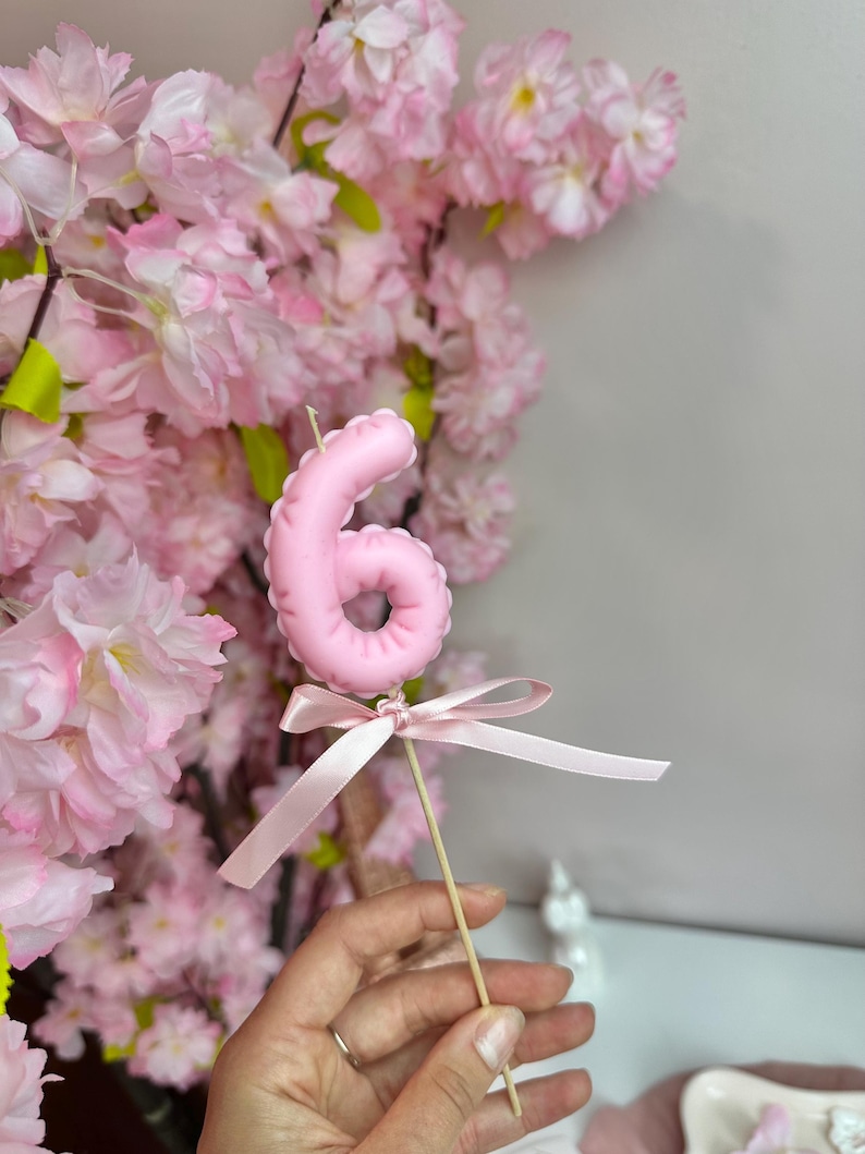 May include: A light pink number six candle topper with a rounded, inflated appearance, featuring a thin satin ribbon tied in a bow at the base of the number, attached to a slender wooden stick for placement in a cake or dessert.