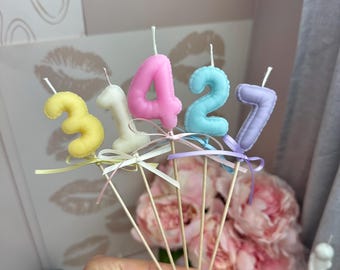 Puede incluir: Cinco velas numéricas de colores pastel con forma de 3, 1, 4, 2 y 7, cada una unida a un palito de madera. Las velas son de color amarillo, blanco, rosa, azul claro y lavanda, y cada una tiene un pequeño lazo a juego en la base.