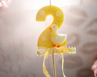 May include: A yellow number two birthday candle featuring a white duck figure, embossed floral patterns, and a light yellow ribbon tied at the base. The candle is mounted on a thin wooden stick, creating a decorative topper for a cake or dessert.