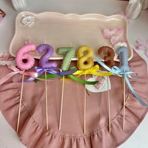 May include: A set of six number-shaped cake candles in pink, purple, green, yellow, brown, and light blue. Each candle is attached to a wooden stick and decorated with a matching satin ribbon bow tied at the base of the number.