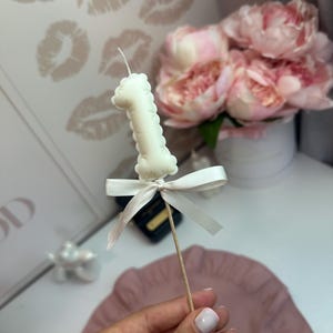 May include: A white candle shaped like the number one, mounted on a thin wooden stick. The candle is decorated with a small, tied white satin ribbon bow just below the number. The candle features a single wick at the top center.