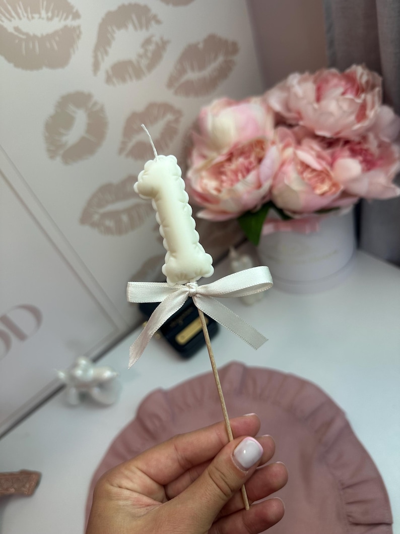 May include: A white candle shaped like the number one, mounted on a thin wooden stick. The candle is decorated with a small, tied white satin ribbon bow just below the number. The candle features a single wick at the top center.