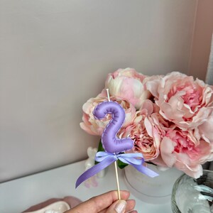 May include: A lavender-colored number two candle shaped like a balloon, featuring a smooth, rounded surface and a white wick at the top. A matching lavender ribbon is tied in a bow around the base of the candle, which is attached to a thin wooden stick.