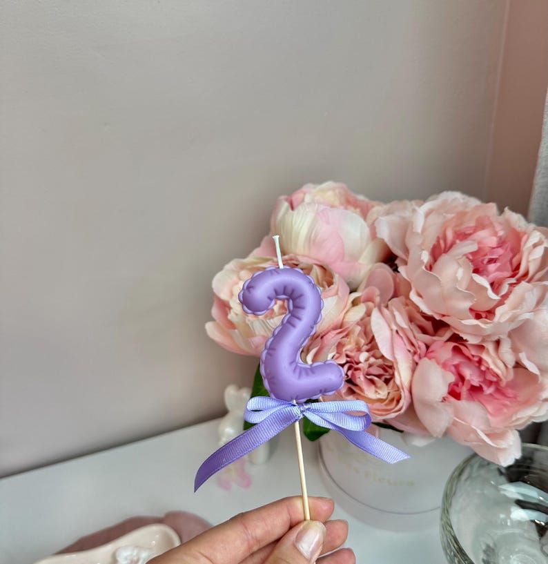 May include: A lavender-colored number two candle shaped like a balloon, featuring a smooth, rounded surface and a white wick at the top. A matching lavender ribbon is tied in a bow around the base of the candle, which is attached to a thin wooden stick.