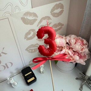 May include: A red number three birthday candle shaped like a balloon, featuring a smooth matte finish and a small red satin ribbon tied around the base of the candle, which is attached to a thin wooden stick for placement on a cake.