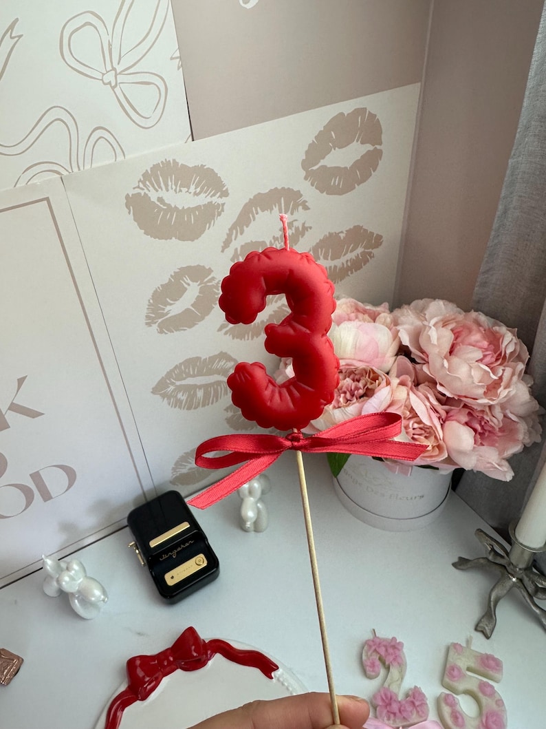 May include: A red number three birthday candle shaped like a balloon, featuring a smooth matte finish and a small red satin ribbon tied around the base of the candle, which is attached to a thin wooden stick for placement on a cake.