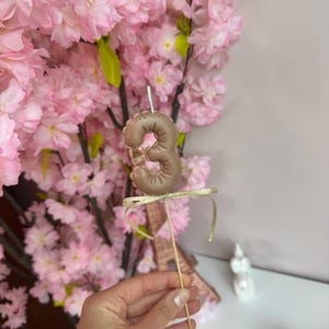 May include: A brown, balloon-style number three cake topper made of a smooth material, featuring a small white wick at the top. The number is attached to a thin wooden stick and decorated with a thin, shimmering gold ribbon tied in a bow.