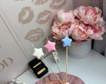 Puede incluir: Tres adornos para velas en forma de estrella en blanco, rosa y azul claro, cada uno unido a un fino palito de madera. Las estrellas tienen un acabado suave y redondeado con una pequeña mecha blanca en la punta superior.