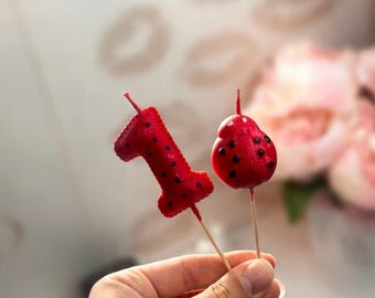 Puede incluir: Dos velas de cera roja sobre palillos de madera. Una vela tiene forma del número uno y la otra forma de mariquita. Ambas velas presentan una superficie texturizada con pequeños puntos negros y una mecha fina en la parte superior.