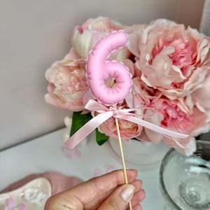 May include: A light pink number six candle topper featuring a smooth, rounded surface. The candle is attached to a thin wooden stick and decorated with a matching light pink satin ribbon tied in a bow at the base of the number.