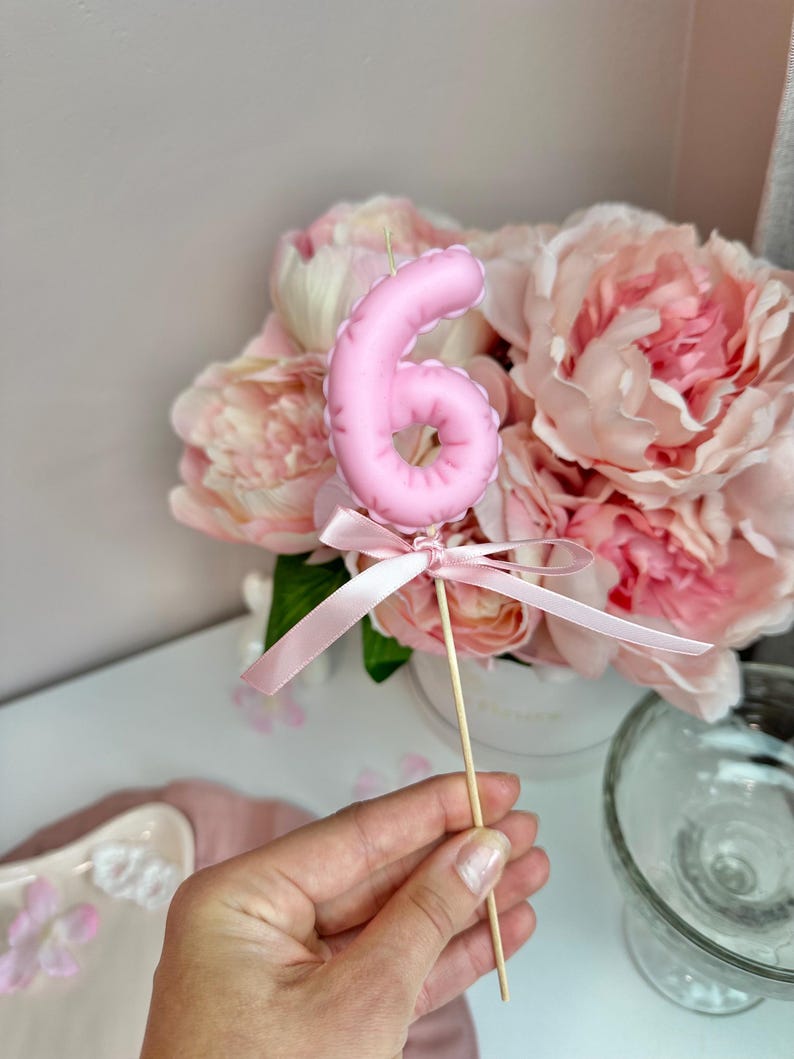 May include: A light pink number six candle topper featuring a smooth, rounded surface. The candle is attached to a thin wooden stick and decorated with a matching light pink satin ribbon tied in a bow at the base of the number.