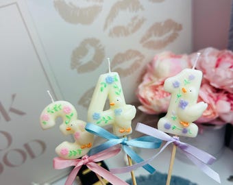 Puede incluir: Tres velas numéricas de color blanco roto con forma de 3, 4 y 1, decoradas con pequeñas figuras de patos moldeadas y motivos florales. Cada vela está sujeta a un palito de madera y adornada con una cinta de raso rosa, azul o lila.