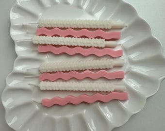 Puede incluir: Un conjunto de ocho velas dispuestas en filas alternas sobre un plato blanco en forma de corazón. Cuatro velas son blancas con una superficie texturizada de cuentas y cuatro velas son de color rosa sólido con forma ondulada.