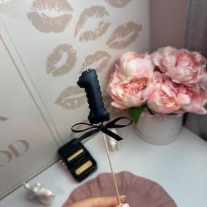 May include: A black number one candle topper featuring a textured surface and a small black ribbon bow tied around the base. The candle is attached to a thin wooden stick, creating a decorative accent for a cake or other food item.