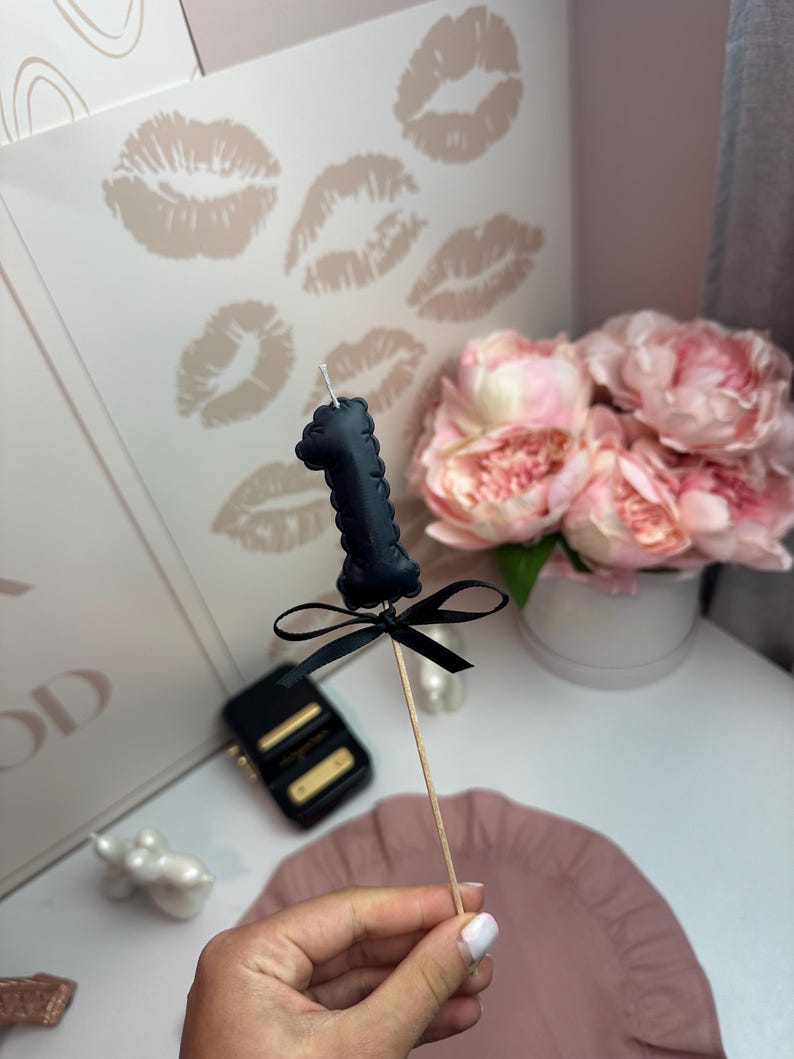 May include: A black number one candle topper featuring a textured surface and a small black ribbon bow tied around the base. The candle is attached to a thin wooden stick, creating a decorative accent for a cake or other food item.