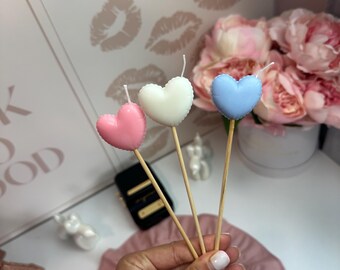 Puede incluir: Tres velas en forma de corazón en rosa pastel, blanco y azul claro, cada una unida a un palo de madera fino. Las velas tienen una superficie lisa y redondeada con una mecha blanca que sobresale del centro superior de cada corazón.