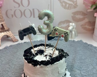 Puede incluir: Un pastel redondo pequeño con glaseado blanco, bordes negros y chispas blancas y negras en la base. Tres adornos están insertados en la parte superior: una vela de color verde claro con el número tres y dos figuras de mandos de juego.