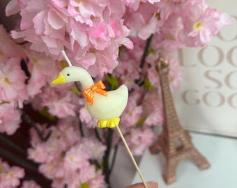 Puede incluir: Una vela con forma de ganso de color crema sobre un palo de madera, con un pico y patas de color amarillo brillante, y un pequeño lazo naranja atado alrededor del cuello. La vela tiene una mecha blanca que sobresale de la cabeza para decoración.