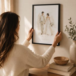 May include: A framed watercolor print of a couple holding hands, dressed in wedding attire. The print has a black frame and is being held by a person. The print has the words "Our Forever" and a date.
