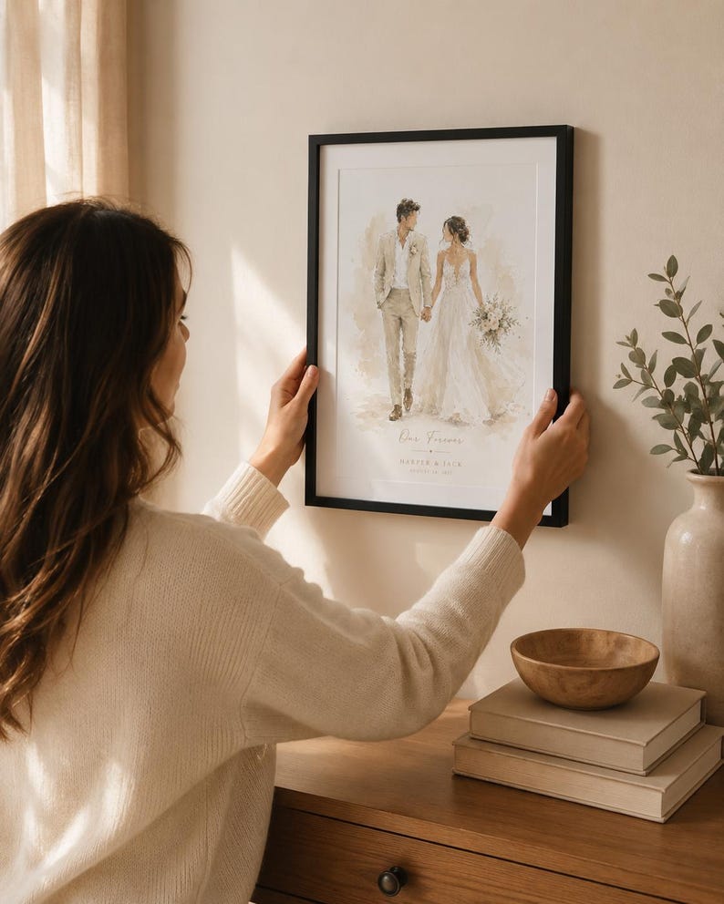 May include: A framed watercolor print of a couple holding hands, dressed in wedding attire. The print has a black frame and is being held by a person. The print has the words "Our Forever" and a date.