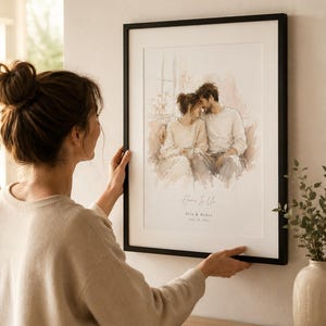 May include: A framed watercolor print with a black frame. The artwork depicts a couple embracing, with soft, neutral tones. The text "Home Is Us" and names are visible. The print is being held by a person.
