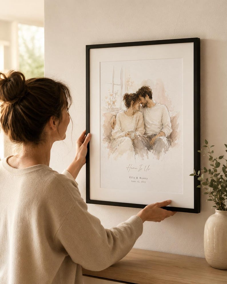 May include: A framed watercolor print with a black frame. The artwork depicts a couple embracing, with soft, neutral tones. The text "Home Is Us" and names are visible. The print is being held by a person.