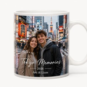 May include: White ceramic mug featuring a photograph of a couple in Tokyo, Japan. The mug has the text "Tokyo Memories 2026 Ava & Liam" in a handwritten style font. The background shows a busy street scene.