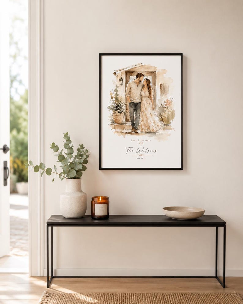 May include: A framed watercolor print of a couple kissing in front of a building. The print is in a black frame and is displayed on a black table with a vase, candle, and bowl. The print has the text "The Wilsons" and "Feb. 2022".