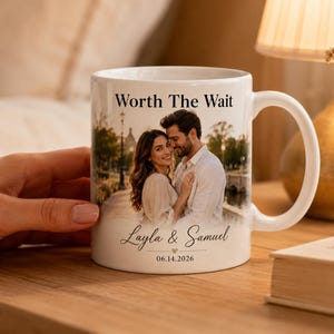 Op de afbeelding: Witte keramische mok met een foto van een stel, de tekst "Worth The Wait" en de namen "Layla & Samuel" met een datum. De mok heeft een gebogen handvat en staat op een houten oppervlak.