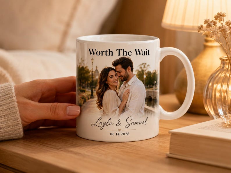 Op de afbeelding: Witte keramische mok met een foto van een stel, de tekst "Worth The Wait" en de namen "Layla & Samuel" met een datum. De mok heeft een gebogen handvat en staat op een houten oppervlak.
