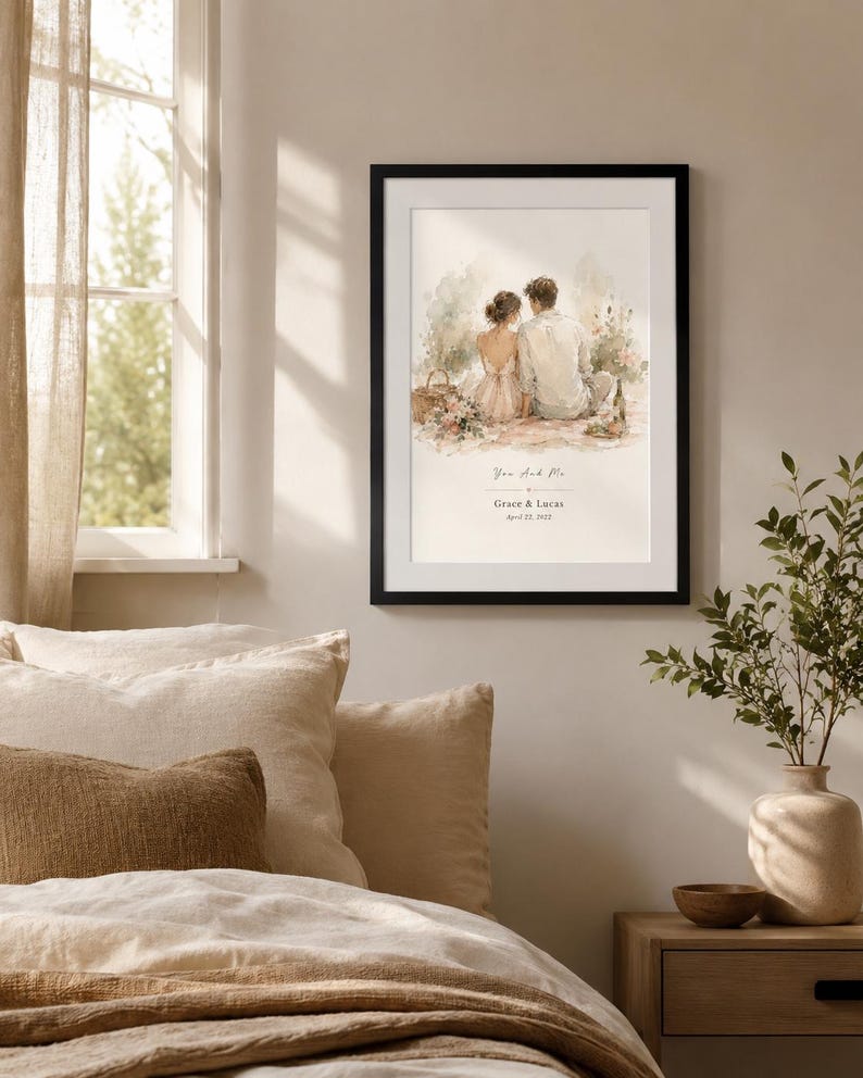 K&ouml;nnte beinhalten: Ein gerahmter Aquarelldruck mit einem Paar, das zusammen sitzt, mit dem Text "You and Me" sowie Namen und Datum darunter. Der Druck befindet sich in einem schwarzen Rahmen und h&auml;ngt an einer neutralfarbenen Wand &uuml;ber einem Bett mit beiger Bettw&auml;sche.