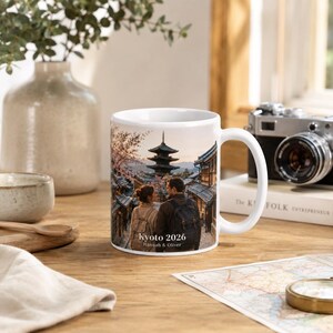 May include: White ceramic mug featuring a scenic image of a Japanese pagoda and street scene. The mug has a curved handle and the text "Kyoto 2026 Hannah & Oliver". The mug is on a wooden surface.