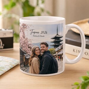 May include: White ceramic mug featuring a photo of a couple in Japan with the text "Japan 2026 Emma & Noah". The mug has a handle and is set on a wooden surface with a camera and map in the background.