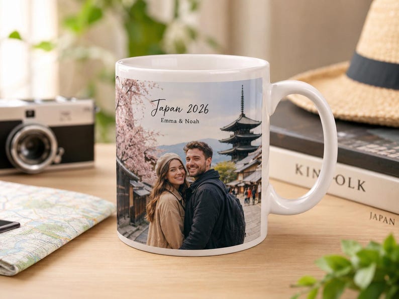 May include: White ceramic mug featuring a photo of a couple in Japan with the text "Japan 2026 Emma & Noah". The mug has a handle and is set on a wooden surface with a camera and map in the background.