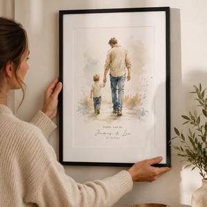 Puede incluir: Una impresi&oacute;n en acuarela enmarcada representa a un padre y un ni&ntilde;o caminando de la mano. La obra de arte presenta azules, beiges y marrones suaves. El texto en la parte inferior dice "Daddy And Me" y "James & Leo" con una fecha. El marco es negro.
