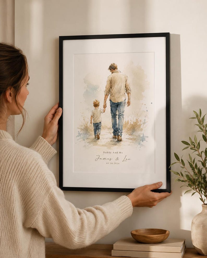 Puede incluir: Una impresi&oacute;n en acuarela enmarcada representa a un padre y un ni&ntilde;o caminando de la mano. La obra de arte presenta azules, beiges y marrones suaves. El texto en la parte inferior dice "Daddy And Me" y "James & Leo" con una fecha. El marco es negro.