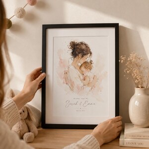 May include: A framed watercolor print depicts a mother and child in soft pink and beige tones. The black frame surrounds the artwork, which includes the text "Mommy and Me" and the names "Sarah & Emma." A decorative piece for a nursery or home.