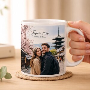 May include: White ceramic mug featuring a couple in front of a Japanese pagoda and cherry blossoms. The mug has the text "Japan 2026" and the names "Emma & Noah". The mug is on a woven coaster.