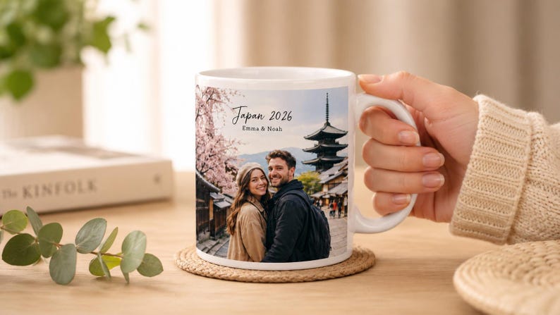 May include: White ceramic mug featuring a couple in front of a Japanese pagoda and cherry blossoms. The mug has the text "Japan 2026" and the names "Emma & Noah". The mug is on a woven coaster.