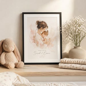 May include: A framed watercolor print featuring a mother and child in soft pink and beige tones. The print is in a black frame and includes the text "Mommy And Me" and the names "Sarah & Emma". A plush bunny and folded blankets are on the shelf.