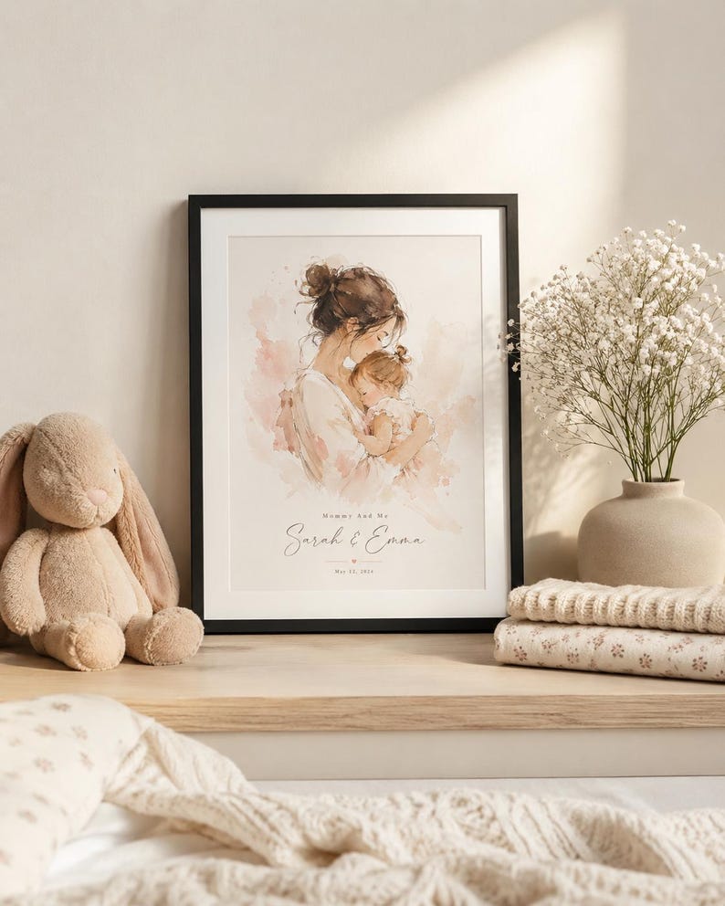 May include: A framed watercolor print featuring a mother and child in soft pink and beige tones. The print is in a black frame and includes the text "Mommy And Me" and the names "Sarah & Emma". A plush bunny and folded blankets are on the shelf.