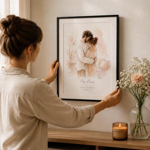 May include: A framed watercolor print featuring a couple embracing, with the text "My Person" and names below. The artwork is in a black frame, displayed on a wall. A woman is shown holding the frame. A vase of flowers and a lit candle are on a wooden surface.