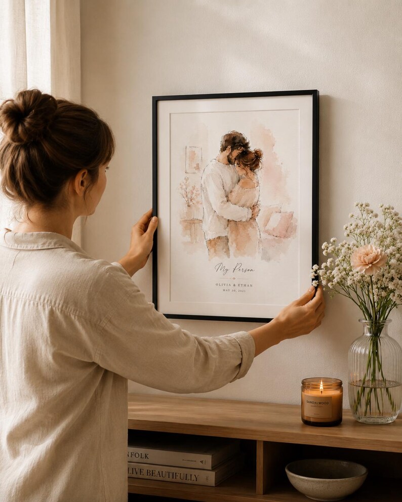 May include: A framed watercolor print featuring a couple embracing, with the text "My Person" and names below. The artwork is in a black frame, displayed on a wall. A woman is shown holding the frame. A vase of flowers and a lit candle are on a wooden surface.