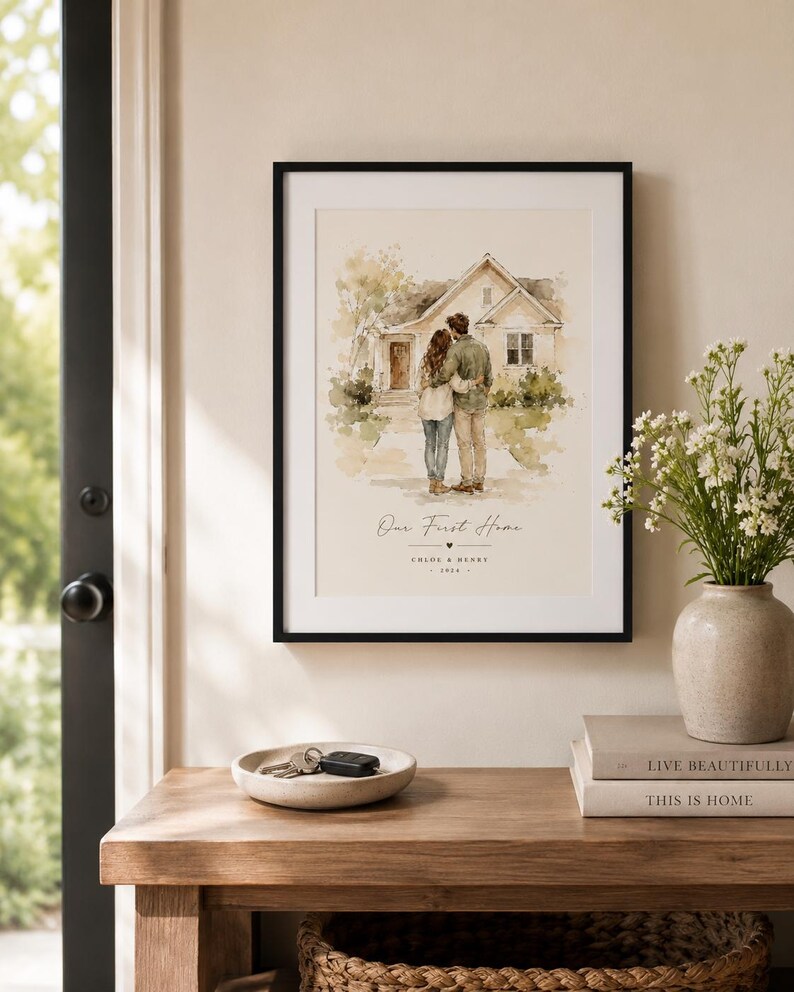 Puede incluir: Una impresi&oacute;n enmarcada con un marco negro, que representa a una pareja abraz&aacute;ndose frente a una casa. La impresi&oacute;n se titula "Our First Home" con nombres y un a&ntilde;o debajo. La impresi&oacute;n se exhibe sobre una mesa de madera con un cuenco decorativo y libros.