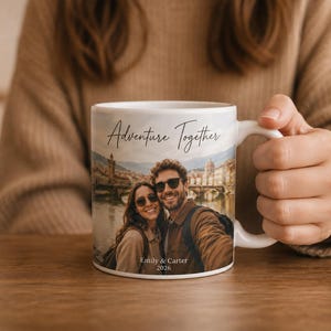 Pu&ograve; includere: Tazza in ceramica bianca con la scritta "Adventure Together" e una foto di una coppia. La foto mostra una coppia davanti a un ponte e degli edifici. La tazza riporta anche i nomi "Emily & Carter" e l'anno "2026".