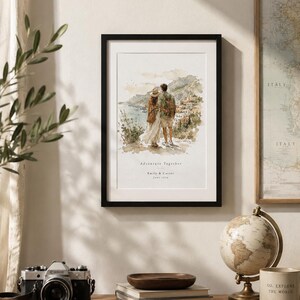 K&ouml;nnte beinhalten: Ein gerahmter Aquarelldruck mit schwarzem Rahmen. Der Druck zeigt ein Paar mit Blick auf eine K&uuml;stenstadt. Der Text "Adventure Together" und Namen stehen darunter. Eine Vintage-Kamera, B&uuml;cher und ein Globus stehen auf einem Holzregal.