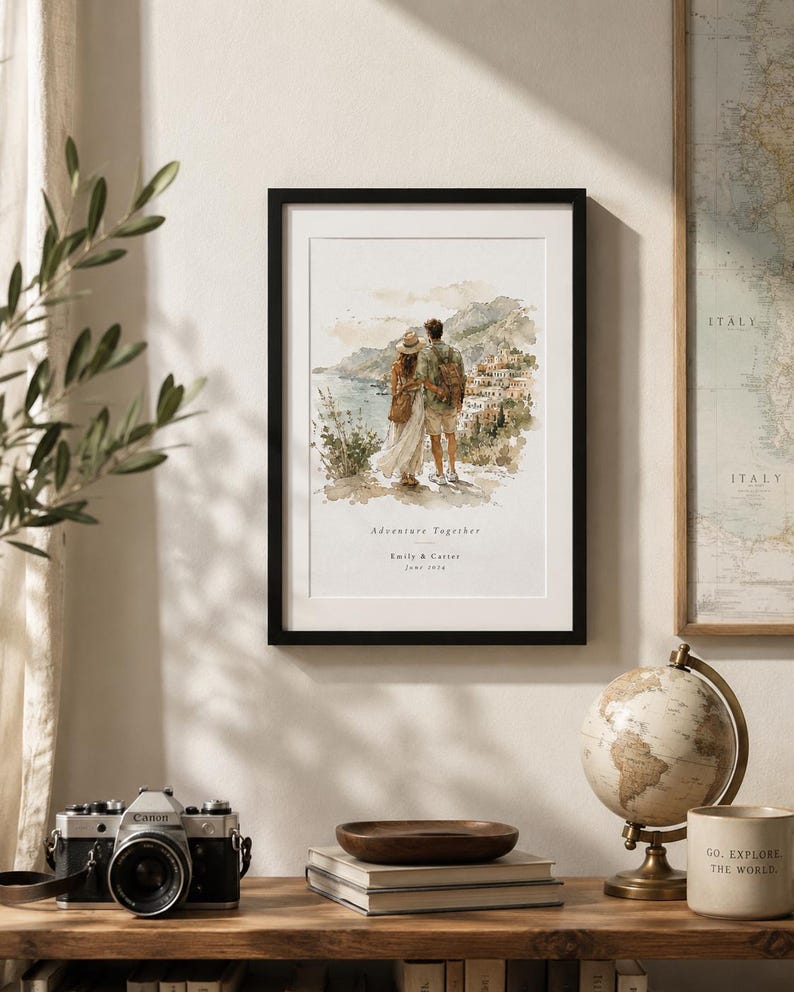 K&ouml;nnte beinhalten: Ein gerahmter Aquarelldruck mit schwarzem Rahmen. Der Druck zeigt ein Paar mit Blick auf eine K&uuml;stenstadt. Der Text "Adventure Together" und Namen stehen darunter. Eine Vintage-Kamera, B&uuml;cher und ein Globus stehen auf einem Holzregal.