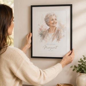 May include: A framed watercolor portrait of a woman with the text "Always in my heart" and the name "Margaret" below. The portrait is in a black frame and is being held up on a wall.