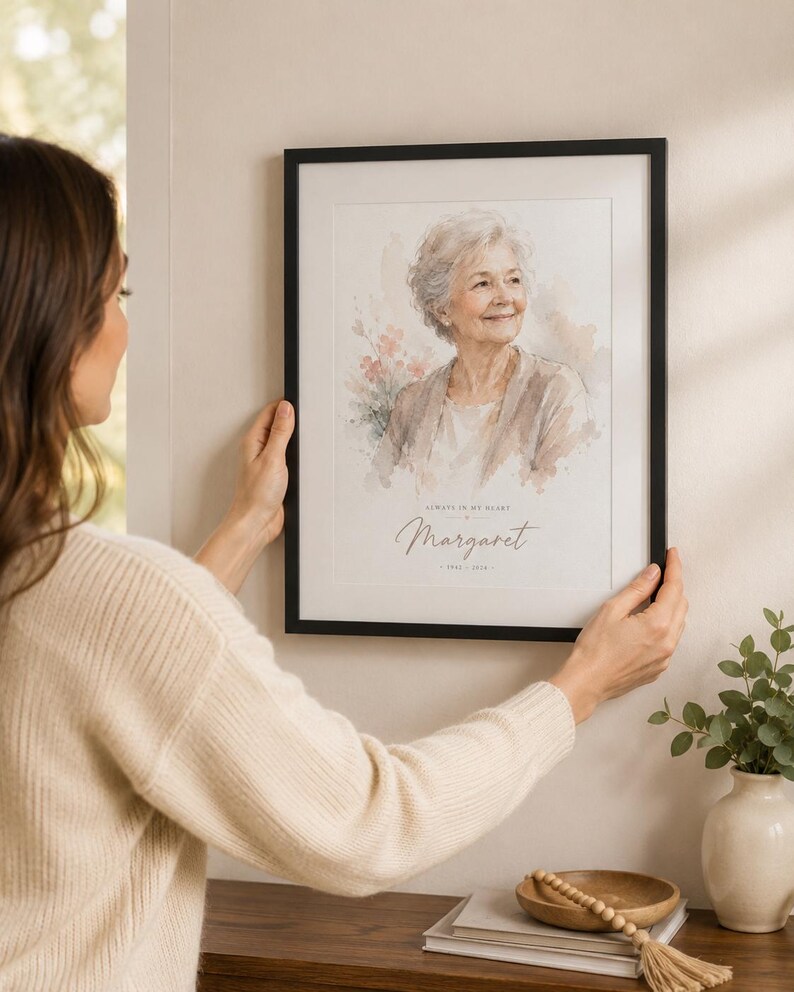 May include: A framed watercolor portrait of a woman with the text "Always in my heart" and the name "Margaret" below. The portrait is in a black frame and is being held up on a wall.